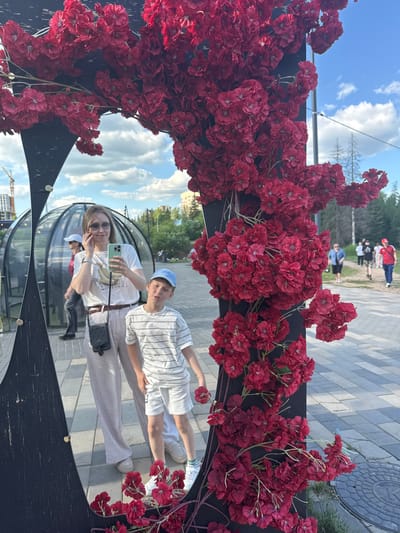 The image captures a woman and a child reflected in a decorative mirror within the Garden of the World (Сад Мира) in Izhevsk, Russia. The mirror features an irregularly shaped dark frame, heavily adorned with clusters of artificial red flowers, consistent with a cherry blossom aesthetic.
In the reflection, a woman with light brown hair, wearing sunglasses, a white t-shirt with a graphic design, light-colored striped pants, and a dark cross-body bag, holds a light green smartphone, appearing to take a photograph or selfie. A metallic watch is visible on her left wrist. Adjacent to her, a young boy wears a light blue baseball cap, a white horizontally striped t-shirt, white shorts, and sneakers. He is making a playful pouting expression and holds a single artificial red flower in his right hand.
Behind the reflected subjects, several transparent, dome-shaped structures, characteristic of geodesic domes, are visible. The immediate ground in the reflection consists of light-colored pavement.
Beyond the mirror's decorative frame, the environment shows an outdoor public space on a clear day with a blue sky and scattered white cumulus clouds. Green foliage and trees are visible in the mid-ground and background. Paved pathways extend into the distance, where several indistinct people are seen walking. On the far left, a construction crane and buildings indicate ongoing urban development, with additional buildings visible in the distant right.