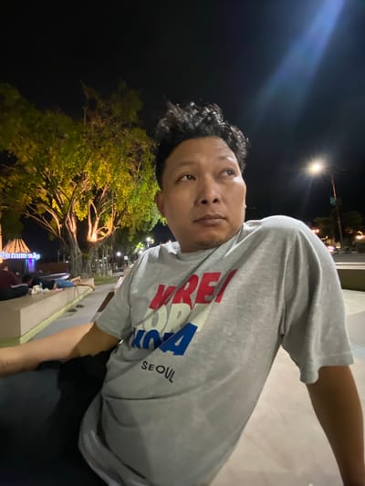 A male individual is captured in a mid-shot photograph taken at night in Sidoarjo, Indonesia, specifically in an area consistent with Monumen Jayandaru. He is of East Asian or Southeast Asian descent, with dark, curly hair and a medium complexion, looking upwards and to his right with a contemplative expression. He wears a light gray short-sleeved t-shirt with the text "KRE" in red, "KO" in white, "REA" in blue, and "SEOUL" in black printed on the front.
The background is an outdoor public space illuminated by various light sources. Large trees with extensive root systems, characteristic of banyan trees, are prominently lit with warm yellow lights on the left, casting a glow on their foliage and trunks. Further left, a large, angular monument structure displays partially visible illuminated text, "Monumen," glowing in blue and purple light. Below the trees, several individuals are visible on a light-colored concrete platform, with at least two lying prone. One individual wears a dark top and light pants, while another wears a red top. A pair of sandals is visible on the platform. On the far right, a bright streetlight casts a prominent lens flare, and distant vehicle lights are faintly visible on a paved surface. The overall scene suggests a vibrant public park or monument area at night.