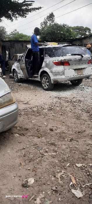 The image depicts an outdoor, unpaved car service area in Nairobi, Kenya. A male individual, dressed in a blue t-shirt and dark trousers, stands with his left foot inside the open driver's door of a silver Honda CR-V-style SUV. He is actively scrubbing the vehicle's roof with a yellow sponge, evidenced by white suds on the roof and rear window. The SUV's rear license plate area is obscured. The ground consists of dirt and gravel, interspersed with various small debris. In the immediate foreground on the left, the front left fender and headlight of another silver vehicle are partially visible. The background features a corrugated metal fence, with dense green foliage and overhead utility wires visible against an overcast sky. Two additional individuals are partially visible: one on the far left behind the foreground vehicle, and another on the far right behind the SUV.