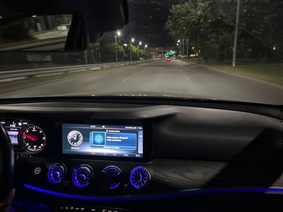 The image shows the interior of a car driving at night on a road in Plovdiv, Bulgaria. The dashboard displays a digital clock showing 00:50 and the date 29.06.2024. The car's infotainment system shows that it is connected to "Daniel's iPhone" and that no Bluetooth audio device is connected. The ambient temperature is 16°C, and the air conditioning is set to 22°C. Outside the car, streetlights illuminate the road, and trees line the sides. Two red taillights of a car ahead are visible in the distance, and traffic lights are also visible. The interior of the car is illuminated by blue ambient lighting.