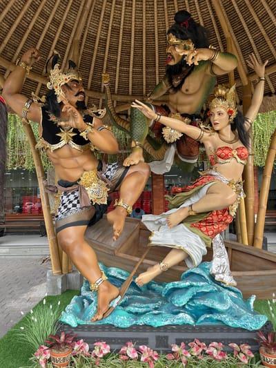 This media file captures a detailed statue display, likely a Balinese cultural representation, set in an outdoor environment. The scene depicts three figures: two male and one female, all rendered with lifelike detail. The figures are adorned in elaborate, traditional costumes, featuring gold accents, intricate patterns, and headdresses. One male figure, positioned lower in the frame, has a muscular physique and wears a checkered sarong and gold jewelry. He appears to be in a dynamic pose, with his mouth open in a roar or shout.
Above him, a green-skinned, reptilian creature with human-like features, including a long tongue, is intertwined with a female figure. This female figure, elegantly dressed in a red and gold top and white skirt, is positioned in a graceful, almost dancing pose, looking upwards with a serene expression. The reptilian creature seems to be holding or interacting with her.
In the background, a representation of turbulent blue water is visible, suggesting a narrative involving a struggle or a mythical scene. The figures are posed on or around structures made of bamboo and wood, with a thatched roof overhead. Green foliage and potted plants surround the display, indicating an outdoor setting, possibly a garden or public space. The overall impression is a vibrant and dramatic portrayal of a mythical or ceremonial event. The lighting suggests daytime, and the presence of plants implies fair weather. No visible text is present in the image. The style and iconography strongly suggest a Balinese origin, possibly from Kuta, Indonesia, given the context.