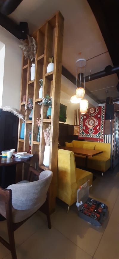 The image shows an interior scene, likely a living room or dining area in Yessentuki, Russia. The room is decorated with a mix of rustic and modern elements. A large wooden bookshelf dominates the left side of the frame, filled with various decorative items, including vases, dried flowers, and what appear to be white ceramic pots.

In the background, a red and beige patterned rug is laid on the floor, near a bright yellow sofa. Above the rug, a modern chandelier with three globe-shaped lights hangs from the ceiling. A black pipe, possibly part of a heating system or chimney, is visible on the left wall, extending upwards.

To the right of the yellow sofa, another seating area is suggested by a gray armchair with a wooden frame. In the far right corner, a toolbox is open, revealing a collection of tools. The lighting suggests it could be daytime, though the exact time is unclear. There are no people visible in the image, and the overall atmosphere appears to be one of a lived-in, cozy space.