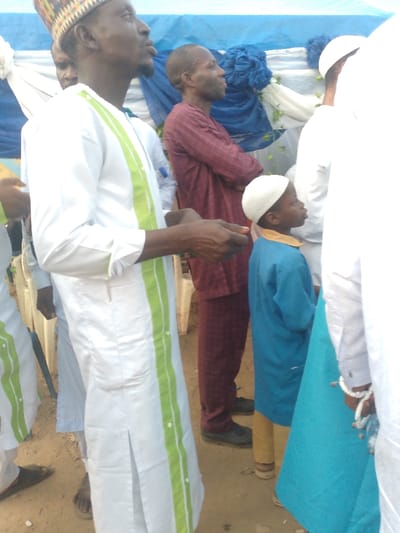 A group of people stands outdoors, dressed in various attire. A tall man in a white robe with green stripes wears a patterned hat, looking upwards. Beside him, a man in a maroon suit stands with arms crossed. A young boy in a blue robe and white cap looks in the same direction. Other individuals are partially visible, some wearing white robes and caps, suggesting a religious or cultural gathering. The backdrop includes draped white and blue fabrics.