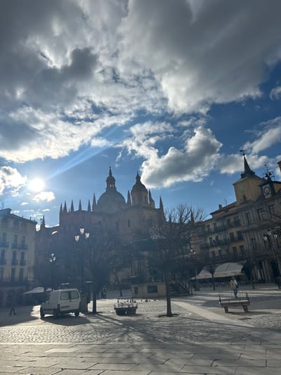 The image shows the Plaza Mayor in Segovia, Spain, located at the coordinates 40.95 latitude and -4.124 longitude. The scene is a square, paved with cobblestones, and surrounded by buildings. In the background, a large church with two towers is visible, partially obscured by trees. The sun is shining brightly in the sky, casting long shadows across the square. A few people are walking in the square. A white van is parked on the left side of the square. There are also several trees in the square, and a bench is visible on the right side of the square. A canopy is seen on the right side of the image, with a few people visible under it. The buildings surrounding the square have a mix of architectural styles, including traditional Spanish and modern. The scene is peaceful and quiet, with a sense of serenity.