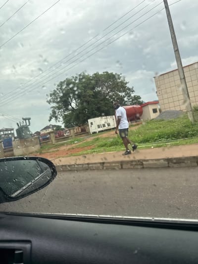 The image, taken from inside a vehicle, shows a view of a street scene in Lugbe, Nigeria. Raindrops are visible on the car window and side mirror. On the right, the edge of the road is visible with a sidewalk adjacent to it. A man wearing a white shirt and dark shorts is walking on the sidewalk. In the background, there are buildings, trees, and electrical power lines.