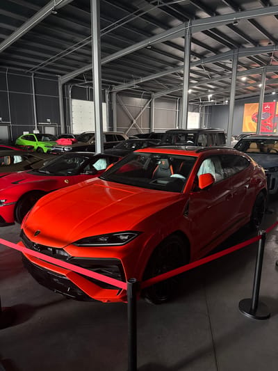 The image shows an indoor exhibition of luxury cars. The primary subject is a bright orange Lamborghini sports car, angled in the foreground. It has black wheels and orange accents visible on the interior. Other cars, including a bright green one and a red one, are parked behind it, also appearing to be high-end vehicles. The setting is a large, industrial-looking building with a high ceiling supported by metal beams and a dark, ribbed ceiling. Industrial lighting is visible. A red ribbon barrier is set up in front of the orange car, suggesting it is part of a display or event. The floor is dark and appears to be concrete. In the background, a large poster featuring a red sports car is hanging on the wall. The overall atmosphere suggests a car show or dealership event. There are no people visible in the image. The location is Plovdiv, Bulgaria.
