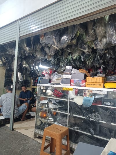 The image captures the interior and immediate exterior of an auto parts shop located in Bogor, Indonesia, viewed from an outdoor paved surface. The shop features an open front with a partially raised metal roll-up security shutter. The interior space is densely packed with automotive components, primarily headlights, taillights, and various plastic body parts, many individually wrapped in clear plastic and suspended from the ceiling or stacked. A glass display cabinet occupies the right foreground, filled with assorted vehicle lights and plastic panels. On top of this cabinet, cardboard boxes are stacked, some labeled "XENON HID" and "VND."
Three adult males are visible within the shop. On the left, one man wearing a grey t-shirt and blue denim shorts is seated on a low platform, largely facing away from the camera. In the center, another man, wearing a dark blue t-shirt and blue shorts, sits on the floor, holding a dark object, and is smiling broadly towards the camera. A third man, partially obscured, is visible deeper within the shop, wearing a dark green cap. A light brown plastic stool is positioned in the foreground on the paved exterior. The shop interior appears dimly lit beyond the entrance, relying on natural light from the open front.