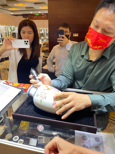 In a brightly lit indoor setting, possibly a shop or exhibition, a group of people are examining items in display cases. The image is oriented vertically.
In the foreground, a woman with long, dark hair is holding a white smartphone and looking at it. She is wearing a white top over a black garment. To her right, a man in a green shirt and a red face mask with gold Chinese characters and "2022" is looking down at a vase. His hands are visible as he holds the vase, which is white with a floral design. Another man, partially visible in the reflection of a display case or window, is wearing glasses and a black mask and holding a smartphone.
The objects of interest include the decorated vase, a laptop on a table, and various items within glass display cases, appearing to be coins or medallions in holders. The display cases are well-lit, showcasing the items within. The background shows more display cases and architectural elements of a building, suggesting a commercial or public space. The overall atmosphere appears to be one of focused examination and perhaps appreciation of the displayed items. No information about the time of day or weather is available. Visible text includes "2022" on the face mask and some unreadable Chinese characters on various labels within the display cases.