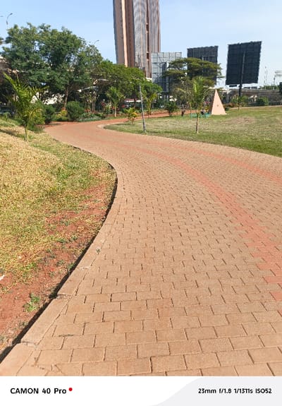A brick pathway curves through a park, lined with grass and trees. A modern, tall building and billboards rise in the background. The path, constructed of tan bricks with red accents, leads the viewer's eye into the scene. The image appears to have been taken on a sunny day. Camera details "CAMON 40 Pro" are at the bottom left, and "23mm f/1.8 1/1311s ISO52" are at the bottom right.