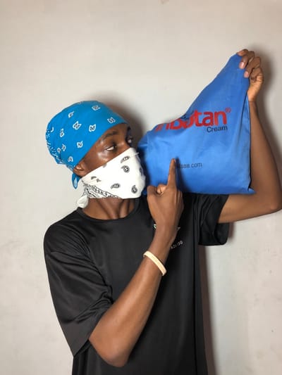 A male individual is depicted from the chest up, positioned against a plain light grey wall in Ngleshi Amanfro, Ghana. He wears a blue bandana with white paisley patterns covering his head and a white bandana with black paisley patterns covering his nose and mouth. His attire includes a black short-sleeved t-shirt with faint text visible on the chest. A light-colored bracelet adorns his right wrist. He holds a blue bag over his right shoulder. The bag features red text reading "inbotan® Cream" and black text displaying "hudson.com". The individual's right index finger is extended, pointing directly at the "hudson.com" text on the bag. His eyes are directed upwards and to the right, consistent with his head's slight tilt.