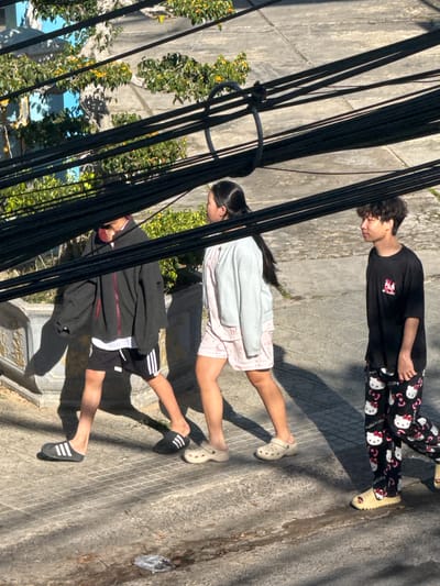 The image shows three young people walking on a paved sidewalk in Da Lat, Vietnam. The weather appears sunny with shadows indicating a midday sun. Overhead, a dense network of black electrical wires spans across the frame.
On the left, a person wearing a black zip-up hoodie, black shorts with white stripes, and black Adidas slides walks with their head down. A portion of a white t-shirt is visible under the hoodie.
In the middle, a person in a light gray zip-up hoodie and pink shorts with a white pattern walks alongside. They are wearing beige Crocs. Their dark hair is pulled back in a ponytail.
On the right, a person with curly dark hair wears a black t-shirt with a graphic print and black pants adorned with a repeating pattern of Hello Kitty faces and bows. They are wearing cream-colored platform slides. Their gaze is directed to the side.
The background consists of a textured gray pavement, a low stone wall with ornate carvings, and some greenery with yellow flowers. The environment suggests an urban or semi-urban setting.
