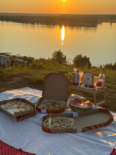 This is a forensic, matter-of-fact description of a picnic scene set against the backdrop of a sunset over a body of water, likely the Volga River, near the city of Olkhovka, Russia.
The scene is set on a grassy slope overlooking a wide river. The sky is ablaze with the warm hues of sunset, casting a golden glow on the water. A sunlit reflection stretches across the river's surface. In the distance, the opposite bank is lined with trees and a hazy horizon. On the near bank, some buildings are visible, suggesting proximity to a settlement.
In the foreground, a white picnic blanket is spread out. On the blanket, there are several pizza boxes. One box is open, revealing a pizza with a visible crust and toppings. Another open box contains a pizza with a single slice missing. A third pizza box is also open and shows a pizza with mushroom toppings. A half of a watermelon, wrapped in plastic, is positioned near the pizzas.
A small portable wooden table is set up. On the table, there are several items: a bottle of pink sparkling wine, a bottle of what appears to be mineral water, three clear plastic cups with pink liquid, two bags of potato chips, and a small plate with three pieces of fruit, likely peaches. A white mobile phone is also visible on the blanket.
The overall scene depicts a leisurely outdoor meal during a sunset. There are no people visible in the image. The environment is natural, with grass and trees, and the presence of the river and distant buildings indicates a semi-rural or edge-of-town setting.