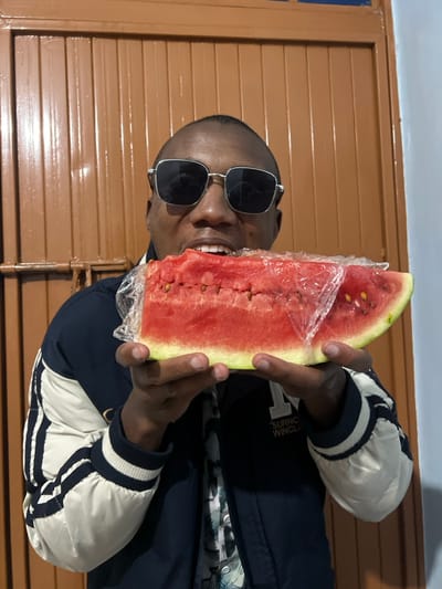 A man wearing sunglasses and a jacket holds a large slice of watermelon wrapped in plastic wrap in front of his mouth, appearing about to take a bite. He is indoors, standing in front of a brown paneled wall. He is smiling and his teeth are visible. The jacket is dark blue with white and black striped sleeves.