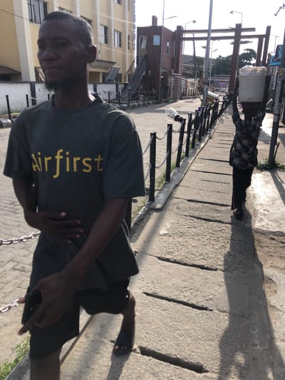 The image presents a man wearing a gray "Airfirst" t-shirt and black shorts, holding a cell phone. He stands on a paved sidewalk with recessed lines, alongside a black chain-link fence. Behind the fence is a building with multiple windows. In the background, another person stands further down the sidewalk, holding what appears to be a bucket or container atop their head, under a metal structure. The location is likely Ado, Nigeria, based on provided context. The overall lighting is bright, casting strong shadows across the pavement.