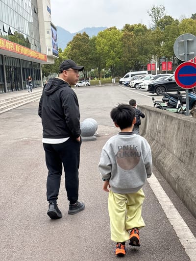 A daylight image depicts an East Asian adult male and two East Asian boys walking on a paved pathway in Jiangjin, China. The adult, approximately 30-40 years old, wears a black baseball cap, black hooded jacket over a white t-shirt, black sweatpants, and black sneakers. He walks with his hands in his pockets, his head slightly turned to the left, and wears eyeglasses. Ahead and to his right, a young boy, approximately 5-8 years old, wears a grey long-sleeved sweatshirt featuring the text "PNIK KUKMU" over a brown circular graphic on the back, light yellow sweatpants, and orange/black sneakers. A second, younger boy in a dark jacket is partially visible, walking further ahead of the first child.

To the left of the pathway stands a modern, multi-story building with a glass facade and white stone base, displaying red banners with Chinese characters. A female figure in a dark jacket is visible ascending its steps in the distance. Two grey spherical stone bollards are positioned near the building's base. To the right, a parking lot contains multiple parked vehicles including white vans, SUVs, and at least two electric scooters, separated from the pathway by a low grey concrete barrier. A red circular traffic sign with a horizontal white bar is prominently displayed on the right. The background features a line of green trees and distant, hazy mountains under an overcast sky.