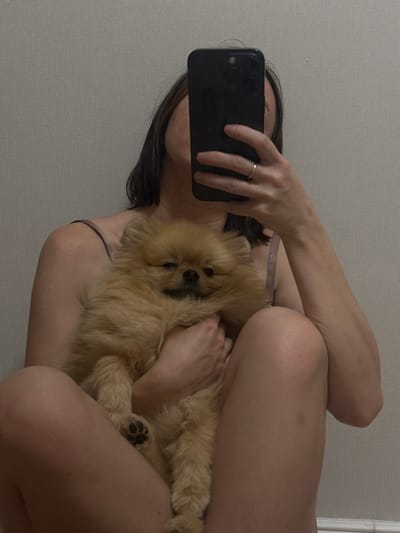 A medium shot captured from a low angle shows a person seated with their legs crossed, holding a small, fluffy, light brown Pomeranian dog. The person is holding a black smartphone up to take the photo, obscuring their face. A thin gold ring is visible on the ring finger of the hand holding the phone. The person is wearing a light-colored camisole. The background is a plain, textured light gray wall. The lighting is soft and appears to be artificial. This image was taken indoors.