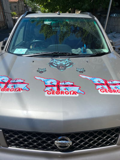 The image shows the hood of a silver car, likely a Nissan given the badge on the grille. The hood is decorated with several stickers. Prominently displayed are three stickers shaped like the map of Georgia, filled with the Georgian flag. The flag features a white background with a large red cross, and four smaller red crosses in each quadrant. The word "GEORGIA" is written in red letters on a white background next to two of these map stickers.

There are also two stickers of wolf heads, one larger and more detailed than the other, which appear to be snarling. The larger wolf sticker is positioned on the left side of the hood, closer to the windshield.

The car's windshield is visible, reflecting trees and the sky. A small blue device, possibly a GPS unit, is attached to the windshield on the passenger side, with a grey microfiber cloth underneath it.

The background suggests an outdoor setting in a city, with hints of buildings and trees visible. The lighting indicates it is daytime, possibly a sunny day given the bright reflections on the car. The presence of the Georgian flag stickers strongly suggests the location is Georgia. The overall impression is one of personal expression and national pride displayed on a vehicle. No people are visible in the image.