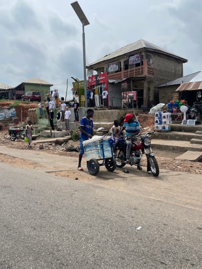 In Lokoja, Nigeria, a street scene unfolds with a man pushing a cart with a blue and white cooler, labeled "Fresco Milk." He is dressed in a blue t-shirt and denim shorts. Seated on a red motorcycle beside him is another individual wearing a striped shirt and a red cap, also in denim. The motorcycle is parked on a dirt road adjacent to a paved area. Behind them, a cluster of mannequins is displayed on raised steps in front of shops. These mannequins are dressed in various casual outfits. The shops appear to be small, local businesses, with signs visible, including "Joe Fashion" and "Sensema's Gadget Store." One building has a multi-story structure with a balcony. A red SUV is parked on the left side of the scene. The environment suggests an outdoor market or commercial area in a town or city, characterized by a mix of shops, display areas, and pedestrian activity. The sky is overcast.