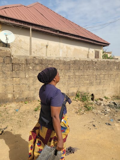 The image depicts a person standing outdoors in Barakin Akawo, Nigeria, under bright daylight. The subject, viewed from the right side in a three-quarter profile, appears to be a woman with dark skin, wearing a dark blue short-sleeved t-shirt, a colorful patterned skirt with red, yellow, and blue designs, and a dark, possibly velvet, head covering. She holds a dark-colored bucket or container in her right hand.

The setting features a dry, sandy, and dusty ground with scattered small rocks, some sparse green vegetation, and debris. Behind the person, a textured light brown wall, possibly made of concrete blocks or stones, runs horizontally. Further into the background, a light-colored building with a distinctive reddish-brown corrugated metal roof is visible against a bright, partly cloudy sky. Electrical wires are strung across the scene, connected to the building. A white satellite dish, emblazoned with the text "SPACE", is mounted on the side of the building towards the lower left. The strong sunlight indicates it is daytime, likely midday or early afternoon, casting clear shadows on the ground. The scene suggests a residential or semi-urban environment.