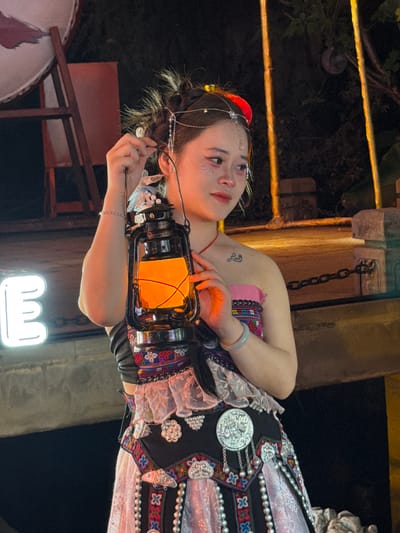 A young woman with elaborate braided hair and metallic headwear stands in what appears to be a nighttime outdoor setting, possibly in Yuzhong District, China. She is holding a vintage-style oil lantern that emits a warm, orange glow. The woman is wearing a traditional outfit with vibrant, embroidered patterns and decorative silver ornaments. Her makeup includes glitter on her eyelids and cheeks. In the background, there is a blurred neon "E" sign to the left and what looks like wooden scaffolding and a large drum to the far left. To the right, there are some illuminated vertical poles and trees, suggesting an outdoor venue or street. A metal chain is visible in the mid-ground.
