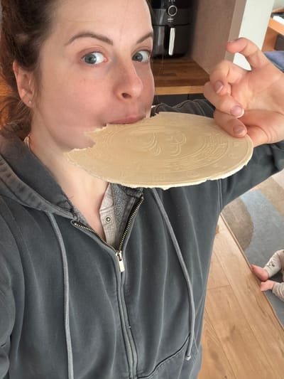 A woman is holding a communion wafer to her mouth, as though about to eat it. She is wearing a gray zip-up hoodie. A baby's feet are visible in the upper right corner of the image. The background is a wooden floor and a kitchen counter with an appliance. This image was taken in Gangloff, Germany.