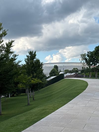 The image depicts a modern, circular building with a colonnade facade, situated in a landscaped park. The building is constructed of light-colored stone and appears to be a stadium or an arena. In the foreground, a paved pathway curves through a manicured lawn. Trees are planted on the lawn, and a hedge runs along the base of an elevated area. The sky is filled with dramatic, dark clouds, with patches of blue sky visible. Two individuals are visible on a stepped area in front of the building, appearing to be sitting and observing. The location is the "Krasnodar Stadium" in Krasnodar, Russia.
