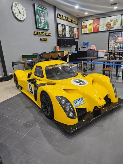 In Na Chom Thian, Thailand, a bright yellow racing car is displayed indoors, positioned on a tiled floor. The car, a prototype with a prominent rear spoiler and aerodynamic bodywork, is the central focus. A large number '9' is visible on the side of the car, and another '9' is on the hood. The car has a license plate with Thai characters and the number '7777'.
Behind the car, the environment appears to be a cafe or restaurant. A large circular clock with Roman numerals is mounted on a grey wall to the left. Above the clock, a green sign with white lettering reads "AI BAAN NAWK COFFEE". Below this, a similar sign in yellow 3D lettering says "AI BAAN NAWK COFFEE". Further to the right, a long panoramic image displays various colorful drinks and desserts. A menu board with Thai text is visible on the wall to the left of the panoramic image. In the background, several tables and chairs are set up, suggesting a dining or seating area. The overall lighting is artificial, with ceiling lights illuminating the space. There are no people visible in the image.