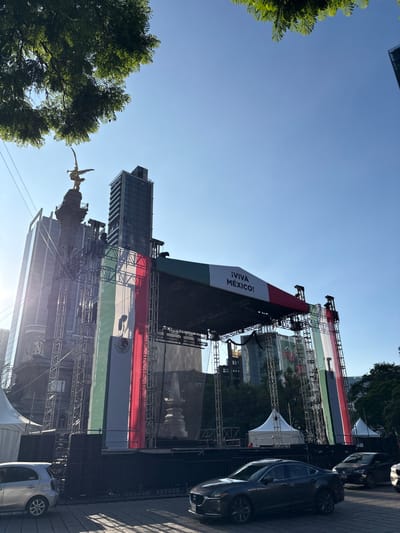 This image depicts a large outdoor stage draped with the Mexican flag and banners, set up in a public space. The Mexican flag is prominently displayed in vertical green, white, and red stripes, with the national coat of arms visible on the white stripe. A large banner with the text "¡VIVA MÉXICO!" is also visible on the stage. The stage is constructed with scaffolding and appears to be part of a celebration or event, likely related to Mexican Independence Day, given the prominent display of national symbols.
In the background, modern skyscrapers rise above the stage, suggesting an urban setting. A golden statue of an angel, likely the Angel of Independence monument, is visible on the left, further confirming the location as Mexico City. Trees with lush green foliage frame the left side of the image, providing a natural element to the urban landscape.
Several cars are parked along the side of the street next to the stage, indicating that the event is accessible by road. The sky is a clear, bright blue, suggesting it is daytime with good weather, likely a sunny afternoon. The overall atmosphere conveyed is festive and patriotic, celebrating Mexican culture and national pride. The image is oriented vertically, with the sky appearing at the top and the ground at the bottom.