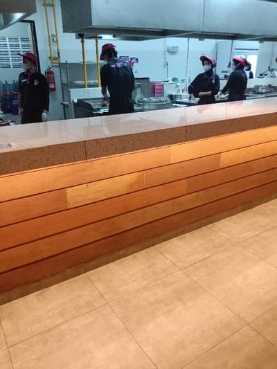 The image shows a view of a kitchen area in a restaurant in Kecamatan Ngaliyan, Semarang, Indonesia. Four people are standing in the kitchen wearing pink hats and face masks.  The kitchen is clean and well-lit, with stainless steel countertops and a wooden wall behind the counter. The counter is made of a light-colored material, likely granite or marble. The floor is tiled with large square tiles.  The wooden wall is made of horizontal planks, likely a type of wood paneling. The people are standing behind the counter and looking towards the left of the frame. There is a large stainless steel hood above the counter, as well as various kitchen appliances. The people are all wearing black shirts, likely employees of the restaurant. There is a large metal container behind the counter.  The image is taken from a low angle, looking up at the counter and wall.  The focus is on the wooden wall and the counter.  The people in the image are blurred, suggesting they are moving or the image is out of focus.  The image is taken in a bright environment, and the lighting is even.  The overall impression is one of a clean, well-maintained kitchen.