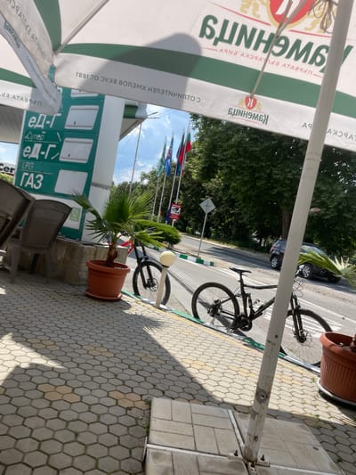 The image depicts a scene outside what appears to be a gas station in Haskovo, Bulgaria. The perspective is from under a large umbrella, looking out onto a sidewalk and a street. A bicycle is parked on the sidewalk next to a potted palm tree. In the background, a dark-colored car is visible on the street, and lush green trees line the roadside. Several flags are also visible in the background.
On the gas station structure, there is prominent signage with Cyrillic text. Some of the visible text includes "SUNHOME," "ед-Г" (which likely refers to LPG gas, given the "LPG" label also present), and "ГАЗ." The text "Cynep" is also visible, possibly indicating a type of fuel or service. The overall atmosphere seems to be a bright, sunny day. There are no people clearly visible in the image, but the presence of the bicycle and the gas station suggests a public outdoor area. The hexagonal paving stones on the sidewalk are a notable detail of the setting. The umbrella overhead casts a shadow on the paving stones.