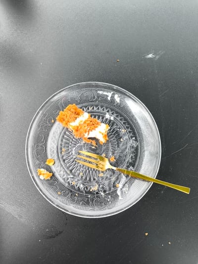 A top-down shot depicts a used clear patterned glass dessert plate on a dark, possibly black, matte surface. The plate contains remnants of a dessert, likely cake, with orange crumbs and white frosting. A gold-colored fork with a bamboo-style handle lies on the plate, partially covered with cake debris. Some cake crumbs are scattered on the surface around the plate. There are no people visible in the image, and the environment appears to be a table setting. The location of the photo cannot be ascertained from the image, and neither can it be confirmed that the picture was taken in Broek op Langedijk, Netherlands.