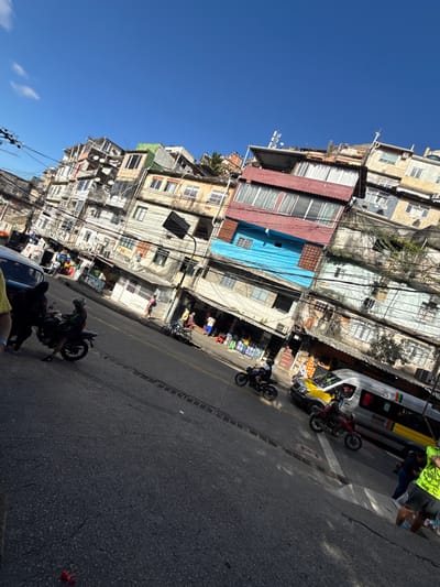 The image depicts a bustling street scene in a densely populated, informal urban settlement characteristic of a *favela* in Rio de Janeiro, Brazil. The photograph is taken from a slightly elevated, oblique angle, looking down a street lined with buildings and under a clear blue sky. The upper portion of the frame is dominated by a bright, cloudless blue sky, with a few faint wispy white clouds visible in the upper left. Below the sky, a dense cluster of multi-story buildings, exhibiting varied construction styles and materials, ascends a hillside. The buildings are painted in a range of colors including white, beige, light blue, reddish-brown, and green. Numerous exposed electrical and communication wires are intricately strung between utility poles and across building facades, forming a dense, chaotic network throughout the midground. Several buildings feature windows, some with protective grates or curtains, and some rooftops display satellite dishes or antennas. The asphalt road, visible in the midground and foreground, is marked by double solid yellow lines. Cracks and wear are evident on the dark grey pavement, and a metal grate or storm drain crosses the road horizontally. A small, crumpled red object lies on the asphalt in the lower-left foreground. Several vehicles and people populate the scene. On the far left, a light-colored car is partially visible. Three motorcycles are in motion or stopped on the road: one dark-colored motorcycle with a rider in a helmet on the left, another dark motorcycle further down the road, and a red motorcycle positioned next to a silver passenger van. This silver van, marked with yellow accents and a multi-colored stripe (red, yellow, green, blue), suggests its use as a public transport vehicle. Multiple individuals are present throughout the frame. In the left foreground, a person in a yellow shirt stands next to the blue car, while another person in dark clothing stands near the dark motorcycle. A rider in a green vest and helmet operates the motorcycle on the left. On the right sidewalk, several pedestrians are visible, including a person in a light pink shirt walking, and another person in a bright yellow-green vest partially visible in the far right foreground. Other people are dispersed along the sidewalks, near storefronts on the right side of the street. These street-level establishments feature open doorways and display various items, with one visible sign bearing the text "JC SUA OBRA". The entire scene is brightly