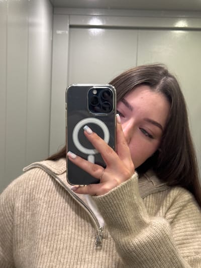 A young woman with long, dark brown hair is taking a selfie in a dimly lit room with beige walls. She is wearing a light beige, ribbed knit sweater with a zippered collar. Her right hand is holding a dark-colored smartphone in front of her face, partially obscuring it. The phone has a clear case with a silver magnetic ring visible on the back. She has manicured nails with a French tip design that fades into light blue. A silver ring is visible on her ring finger. The background appears to be an elevator interior with paneled walls. The lighting suggests an indoor, possibly urban, environment.
