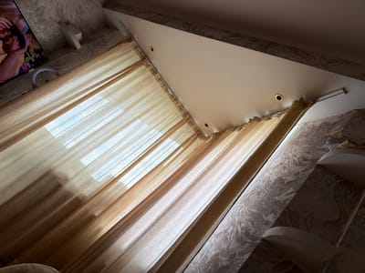 The image depicts an interior scene, likely a room, with a sloped ceiling and patterned wallpaper. A translucent golden curtain is drawn across what appears to be a window, allowing natural light to filter through. The curtain is held by a rail system attached to the ceiling. A portion of a television screen is visible in the upper left corner, displaying an image of people. A ring light is positioned below the television. The overall impression is of a domestic setting. The provided information states the location is Kyiv, Ukraine, but this cannot be visually confirmed from the image itself.
