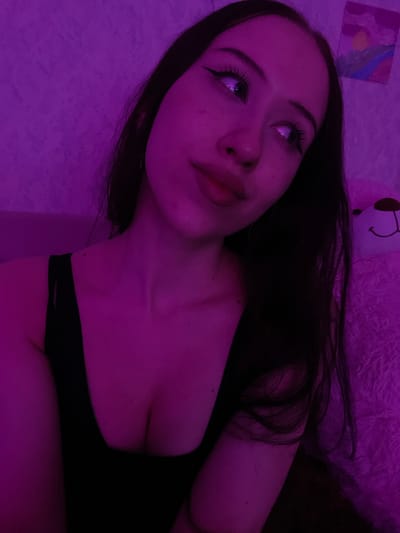 A young woman with long, dark hair and prominent eye makeup is captured in a low-angle, close-up shot, illuminated by purple lighting. She is wearing a black, sleeveless top. Her gaze is directed upwards and to her right. A stuffed bear with a smiley face is partially visible to her right, and a small painting hangs on the wall behind her. The background appears to be a textured wall. The location is Votkinsk, Russia.