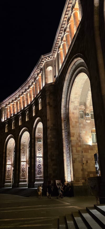 The image depicts a nighttime view of the Republic Square in Yerevan, Armenia, focusing on the northern facade of the National Gallery and History Museum. The building's architecture, characterized by arches and columns, is dramatically illuminated by warm-toned lights. Several people are visible in the foreground, some walking and others standing. The ground area features patterned paving stones and a set of stairs. The sky is dark, indicating it is night.