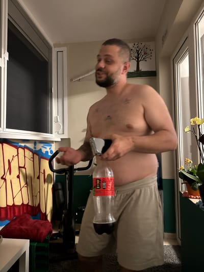 The image shows a bare-chested man holding a plastic bottle of Coca-Cola and a smartphone. He is wearing light-colored shorts. Behind him, there is a bicycle, a wall with graffiti, a window with blinds, and a painting of a tree. The scene appears to be indoors, possibly in an apartment balcony or a room with a view. The presence of a bicycle suggests a fitness area, and the graffiti and art indicate a casual or artistic living space. Without additional context, it is impossible to determine the specific location within the city of Plovdiv, Bulgaria.