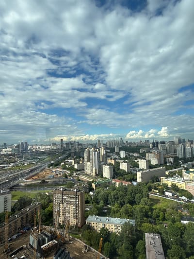 This media file captures an aerial view of a densely populated urban environment with a clear sky and scattered clouds. The scene is dominated by a vast expanse of apartment buildings of varying heights and architectural styles, interspersed with pockets of lush green trees. The perspective suggests a high vantage point, possibly from a tall building or aircraft, looking down and across the city.

In the foreground, the image shows a construction site with multiple cranes and piles of building materials, indicating ongoing development. A complex network of roads and what appear to be railway lines dissects the cityscape, suggesting a major transportation hub. The buildings are mostly residential, characterized by repetitive window patterns and a mix of lighter and darker facades.

The time of day appears to be daytime, with ample natural light illuminating the scene. The weather seems fair, with a partly cloudy sky that allows for good visibility. The presence of numerous trees within the urban fabric adds a natural element to the otherwise built environment. While no people are clearly visible, the extensive infrastructure and housing suggest a significant population. The location is identified as Khimki, Russia, providing a specific geographical context to the urban landscape. There is no visible text in the image.
