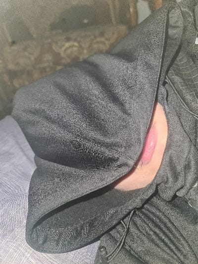 A close-up depicts an open human mouth, partially obscured by draped black fabric. The lips are visible, revealing the inner oral cavity. The fabric forms folds and shadows, adding depth to the composition. A lighter, patterned fabric peeks out from beneath the dark material on one side. The background is blurred, possibly showing elements of an outdoor setting.