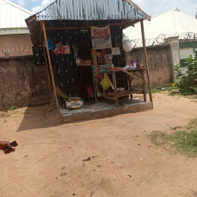 A small outdoor kiosk, likely a convenience store or mobile money agent, is the central subject of this image. The kiosk has a corrugated metal roof and black corrugated metal walls. It is stocked with various goods, including bags of snacks, drinks, and what appear to be eggs and other food items. A POS (Point of Sale) sign is prominently displayed, indicating financial services are offered. A young child, wearing a pink top and a patterned skirt, can be seen inside the kiosk, partially obscured.
The kiosk is situated on a dusty, sandy ground, with a concrete step leading up to it. To the left of the kiosk, a box with "Cavatto" branding is visible. A broom leans against the structure. In the background, a concrete wall topped with barbed wire security fencing runs horizontally, suggesting a built-up area. Beyond the fence, the roof of a modern building with a white, peaked roof is visible, contrasting with the more rustic kiosk. Green foliage can be seen on the right side of the image. The sky is cloudy, suggesting it is daytime, though the specific time is not discernible. The overall scene appears to be a typical street-side establishment in a Nigerian town or city. A hand holding a dark-colored mobile phone is visible in the lower left corner, intruding into the frame from outside the main scene.