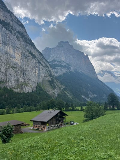 The image depicts a scenic view of the Lauterbrunnen Valley in Switzerland, characterized by towering, steep rock faces and lush green meadows. In the foreground, a traditional wooden barn with a dark metal roof sits in a verdant field dotted with wildflowers. Adjacent to the barn is a smaller structure covered by a dark roof, with a pile of firewood stacked next to it. Two vehicles, a dark-colored car and a white SUV, are parked near the barn. The middle ground is a sloping green pasture, and the background is dominated by immense, craggy mountains, their sheer rock walls textured and streaked with light and shadow. Patches of dark green conifer trees cling to the lower slopes of the mountains. The sky above is a mix of bright blue patches and heavy, grey clouds, suggesting a dynamic weather day. The overall scene conveys a sense of natural grandeur and rural tranquility.