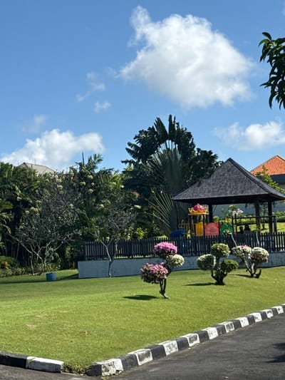 A bright, sunny day in Kuta Selatan, Indonesia, shows a well-maintained green lawn with several manicured bushes and trees. In the background, a dark-roofed gazebo stands next to a colorful playground structure, enclosed by a wooden fence. Beyond the fence, dense tropical foliage and a glimpse of a building with an orange-tiled roof are visible. A paved pathway with black and white striped borders runs along the bottom of the frame. The sky is a vibrant blue with scattered white clouds.