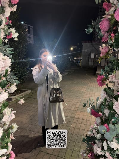 The image captures a young woman at night in what appears to be an urban setting in Batumi, Georgia. The woman, positioned centrally, is taking a selfie or video with her smartphone, which emits a bright light illuminating her face. She has brown hair and is wearing a long, light-colored coat or trench coat and a metallic or silver shoulder bag with a chain strap.

The scene is framed by decorative artificial flowers and foliage in shades of pink, white, and green, suggesting a thoughtfully adorned outdoor space. The ground beneath her is a paved surface with a brick or cobblestone pattern. In the background, under the dark night sky, outlines of buildings and distant artificial lights are visible, contributing to the evening ambiance. To the right of the woman, on the paved ground, there is a distinct black and white QR code printed on a white sticker. The overall mood is casual and modern, characteristic of a night out in a city.