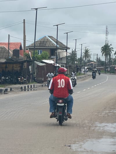 The image depicts a street scene in Badagry, Nigeria, under an overcast sky. A person wearing a red jersey with the number "10" in white, blue jeans, and brown shoes is riding a motorcycle from a rear perspective. They are positioned in the foreground, dominating the center of the frame. The motorcycle has a license plate with a crest.
The road appears to be paved, with a white dashed line indicating the lane. Several other motorcycles and riders are visible further down the road, some moving in the same direction and others in the opposite. To the left of the road, there are buildings with corrugated iron roofs, some appearing to be informal structures or shops, with a water tank visible. Beyond these structures, more distant buildings and vegetation, including palm trees, are present. Utility poles and electrical wires crisscross the sky. In the far background, a telecommunications tower can be seen. The overall environment suggests a bustling urban or semi-urban area.