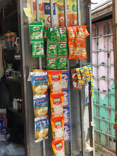 The image displays a detailed street-side retail display within Ngleshi Amanfro, Ghana. The scene centers on a vertical metal grille acting as a merchandise rack, laden with various single-serving packaged goods.
On the upper left, two large green Milo sachets, labeled "No Need to Add Milk," are prominently hung. To their right, two columns each contain four smaller green Milo sachets, marked "Energy to Go Further." Adjacent to these on the upper right, two rows of orange Kivo creamer sachets are suspended.
Below the Milo sachets on the left, three blue and silver sachets of "Cereal Legume Mix," depicting a bowl of cereal, are vertically arranged. Directly to their right, two columns of red and white Kivo "Gari Soaking Mix 4-in-1" sachets are displayed. Further right along the grille, individual sachets of brown Cowbell Coffee, yellow and blue Blue Band margarine, and red and blue "President" branded items are hung.
Behind the metal grille, faded advertisements for mobile network providers such as tiGo, Airtel, Vodafone, Glo, and el are visible. A yellow sticky note is affixed to a metal bar on the upper left.
To the left of the grille, shelving units are partially visible, stocked with items including stacked multi-colored mugs or cups, various plastic bottles, rolls of tissue paper, and transparent plastic bags. A dark bag rests on the ground at the bottom left.
The area to the right of the display grille reveals a corrugated metal wall or barrier, painted in patches of green and a reddish-pink hue, exhibiting paint chipping and signs of wear. The scene is illuminated by natural daylight. No individuals are present in the frame.