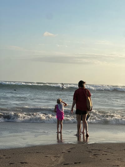In North Kuta, Indonesia, on a beach with wet sand and gentle waves, three people are positioned facing the ocean. A young girl in a pink dress and a white scarf stands in the foreground on the left, looking out at the water. To her right, an adult wearing a red shirt and black shorts is also facing the sea, carrying a large beige bag over their shoulder. Another person, partially visible between the adult and the girl, stands behind them. The ocean has moderately sized waves, and two surfers are visible in the distance. One surfer is riding a wave on the right side of the frame, while another is further out on the left. The sky is a pale blue with a few wispy clouds, and the sun appears to be setting, casting a warm light on the scene.