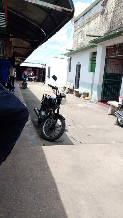 This outdoor scene in Tinaquillo, Venezuela, depicts a bustling street or market area under bright, sunny daylight.
**Subjects:** In the foreground on the left, a person is partially visible, dressed in a dark blue shirt and dark pants. Further into the scene, under a large canopy, several other individuals can be seen walking or standing, though their details are indistinct. The most prominent object is a black motorcycle with a round headlight, parked in the mid-ground on a concrete path. Another blue motorcycle is visible further down the path, and a third motorcycle is partially seen on the far right.
**Scene and Setting:** The left side of the image is dominated by a dark, somewhat rusty metal canopy structure, likely shading market stalls, with glimpses of clothing items hanging. To the right, white-painted buildings with green window frames and metal security grates line the path. These structures exhibit signs of age and wear, including peeling paint and grime. Several light-colored concrete benches are placed outside these buildings. Electrical wires are visible against the cloudy sky above the buildings. The ground is a concrete path, showing varied textures and shadows.
**Activity or event:** The presence of people, market stalls, and parked motorcycles suggests a typical daily scene in a commercial or residential street, with pedestrian traffic and parked vehicles indicating routine activity.
**Notable details:** The strong shadows cast on the concrete path indicate a bright, sunny day, likely mid-day or early afternoon. The sky shows patches of blue with scattered white clouds. The architecture, market setup, and prevalent motorcycles are characteristic of many Latin American towns.
**Visible text:** No legible text is visible on any signs, buildings, or objects in the image.