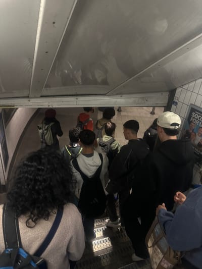 This image captures an interior scene, likely within a subway car or station in London, United Kingdom, depicting a group of people standing. The perspective is from slightly behind and to the right of an individual with dark, curly hair, who is wearing a light grey textured top with a dark strap across their shoulder.
In the middle ground, several other individuals are visible. One person wears a white baseball cap backward and a dark hoodie, looking downwards. Beside them, another person with short dark hair is seen from the side, also appearing to look down. Further into the background, more people are standing, dressed in casual clothing including hoodies and jackets, some carrying bags or backpacks.
To the left of the frame, a large reflective surface or window shows faint, distorted reflections of people and the surroundings. On the right, a wall composed of white rectangular tiles features a poster. Visible text on the poster includes "SUNDAY 23" and "SOCCER FOR U". Below this, a smaller sign displays what appear to be Asian characters. The floor has a distinct, possibly patterned or grated texture. The scene suggests passengers riding or waiting for public transport in an artificially lit indoor environment. No specific emotions are discernible from the individuals.