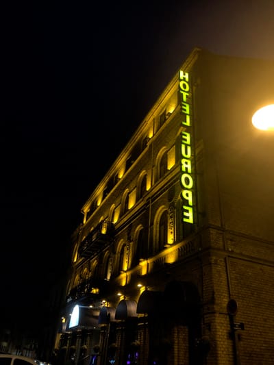 A night view of Hotel Europe, showcasing its architectural details and illuminated sign. The brick facade is lit with warm yellow light, accentuating arches and recesses. The hotel's name, "HOTEL EUROPE," is displayed in bright green neon letters. The image is taken from a low angle, creating a dramatic perspective. A streetlamp above casts a soft glow, while the surrounding darkness enhances the hotel's presence.