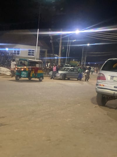 A wide shot captured at night in Gwong, Nigeria, shows a street scene with a few vehicles and pedestrians. In the foreground, a light-colored car is partially visible on the right. To the left, a three-wheeled auto-rickshaw, painted teal and yellow, is the central focus, displaying the text "PTS 2176" on its side. Several people are gathered around the vehicles and in the background, some appearing to be conversing or standing around. The environment is a paved road illuminated by streetlights and possibly car headlights, with buildings visible on the left side of the frame. The overall impression is of a bustling street at night, likely a public area where people are congregating.